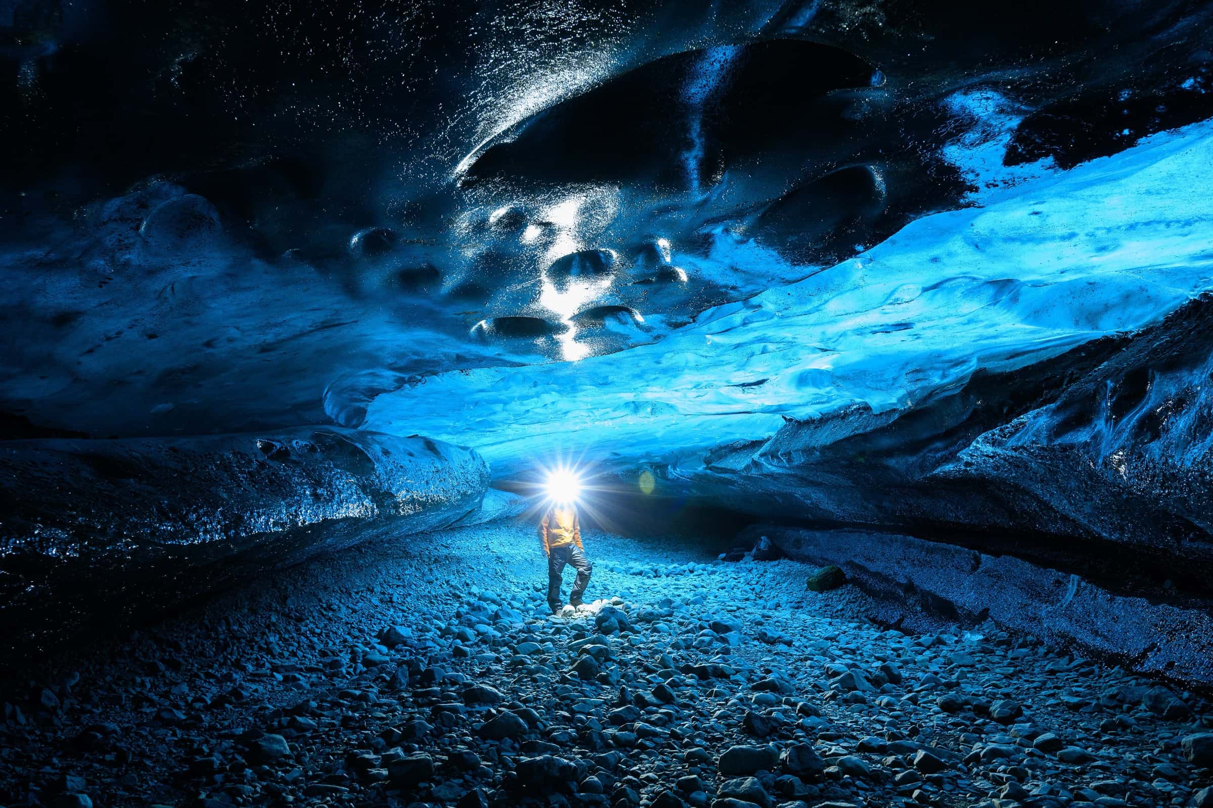 An explorer with a headlamp standing inside a glowing blue glacial ice cave