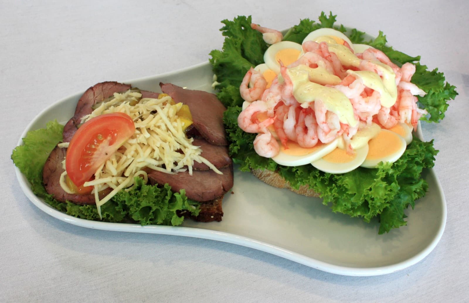 Two smørrebrød open sandwiches plated with roast beef, shrimp, egg and salad